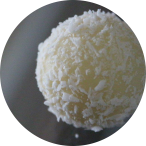 Eine Schokoladenkugel mit weißer Schokolade und Kokosraspeln auf einem grauen Hintergrund. Der Link auf dem Bild führt zur Produktübersicht unserer Confiserie-Produkte.